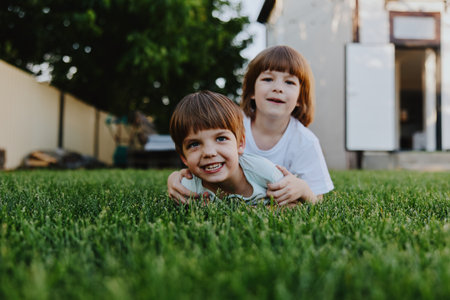Two joyful children playing on green grass, one smiling boy enjoying the moment. A fun and carefree atmosphere in a backyard setting.の写真素材