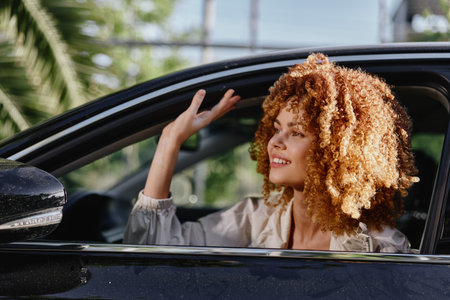 happy woman with curly hair sitting in car window waving hand outdoors on sunny day with green palm...