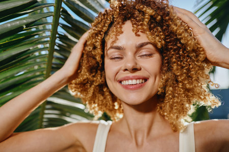 Smiling woman with curly hair enjoys sunlight outdoors near green palm leaves, expressing happiness and natural beauty in warm daylight.の写真素材