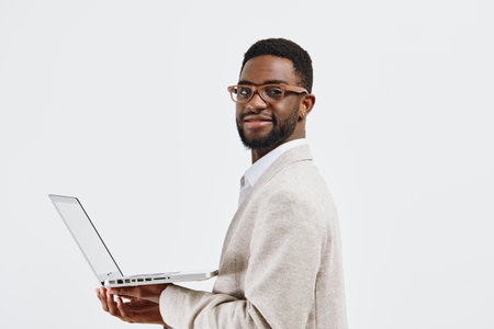 Professional smiling man with glasses, dark curly hair and beard, wearing light-colored blazer and white shirt, holding open laptop, looking at camera on plain white background, studio portrait, business technology conceptの写真素材