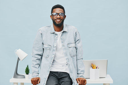 Confident young man with glasses and a light denim jacket sitting in modern office environment smiling, casual clothing, bright background, interior decor, lifestyle concept, positive mood, contemporary styleの写真素材