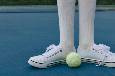 Young athlete standing on a tennis court with white sneakers and knee high socks, focused on the bright green tennis ball, showcasing sport and determinationの写真素材