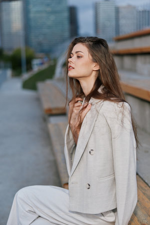 Woman in beige suit sitting on urban bench with pensive expression, natural makeup, and modern office background, stylish fashion portrait outdoorsの写真素材
