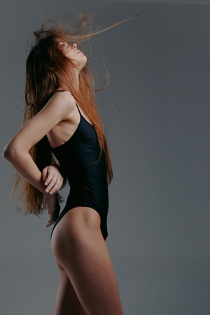 woman in black swimsuit posing with long red hair flowing against gray background in studio...