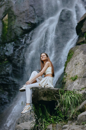 young woman sitting near cascading waterfall outdoors wearing casual white clothes, relaxed and enjoying nature with lush green plants around rocksの写真素材