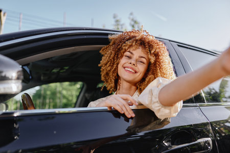 Smiling woman with curly hair leaning out of black car window enjoying sunny day outdoors with...