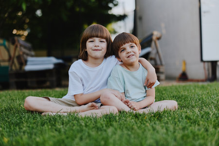 Two smiling boys sitting together on the grass, enjoying their time outdoors with a warm, sunny atmosphere and playful expressions.の写真素材
