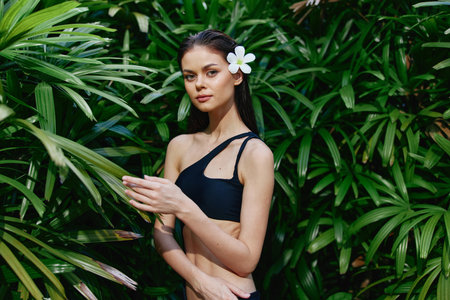Beautiful woman in black swimsuit standing in front of green plants with flower in her hair Summer beauty conceptの写真素材