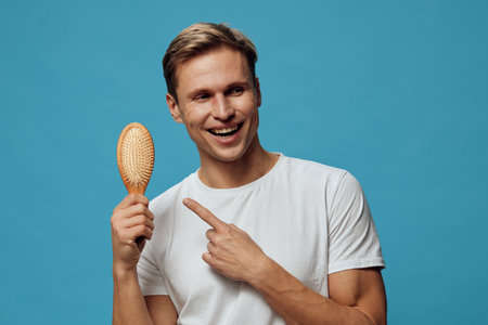 Happy smiling young man with short brown hair wearing plain white t-shirt holding a hairbrush isolated on bright blue background. People lifestyle conceptの写真素材