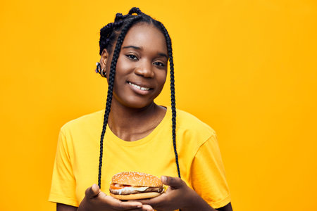 Happy young Black woman holding a delicious burger against a vibrant yellow background, embodying...