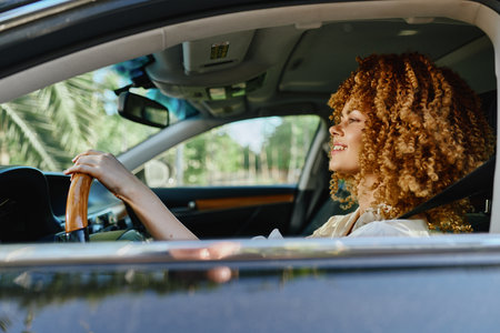 smiling woman driving a car in bright sunlight with natural curly hair and casual beige outfit, enjoying a peaceful moment during daytime.の写真素材