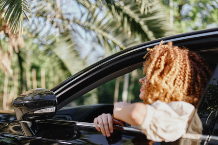 Woman with curly hair looking out of car window, surrounded by tropical palm trees, natural sunlight, and a relaxed summer atmosphere.の写真素材