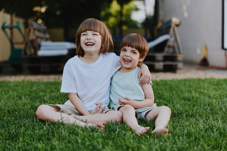 Two happy boys sitting on green grass, sharing laughter and joy. They wear casual summer clothes and have bright smiles, showcasing a playful and carefree moment.の写真素材