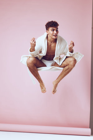 Happy young man in white bathrobe jumping in air with playful expression, against soft pink background. Fun and joy concept in modern lifestyle.の写真素材