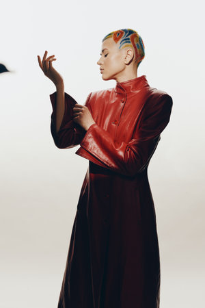 Creative woman with colorful shaved hairstyle wearing a shiny red leather coat, posing thoughtfully against a plain white background in a minimalist studio setting.の写真素材