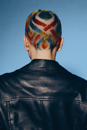 Back view of a woman with colorful geometric shaved hairstyle wearing a black leather jacket, standing against a solid blue background, showcasing bold fashion and creativity.の写真素材