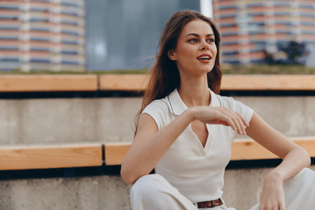 Confident woman with brown hair smiling outdoors in casual white outfit with urban background, natural light, and modern lifestyle conceptの写真素材