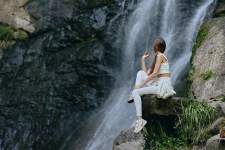 young woman sitting near waterfall in casual white outfit, relaxed, nature, outdoors, adventure, peaceful momentの写真素材