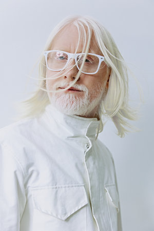 Senior man with long white hair and glasses in a white outfit, showcasing a modern, minimalist style against a soft, light background A portrait emphasizing age and sophisticationの写真素材