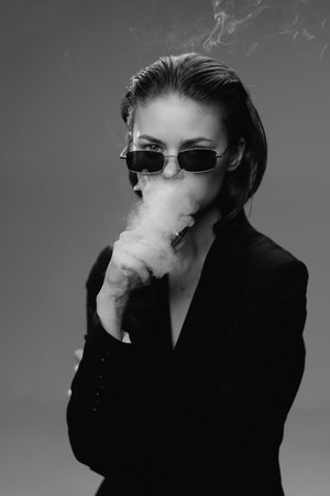 Woman blowing smoke wearing sunglasses in black and white with sleek hairstyle and dark clothing, creating mysterious and stylish atmosphere in monochrome portrait.の写真素材