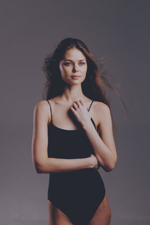 Young woman in black swimsuit poses confidently with minimal makeup and natural hair against a neutral studio background, exuding grace and calm expression.の写真素材