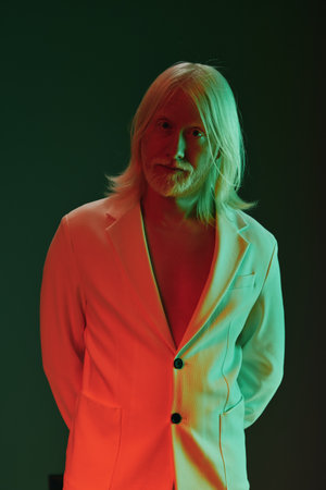 Stylish man with long blonde hair in a white suit against a colorful backdrop showcasing green and red lighting, exuding a confident and artistic vibeの写真素材