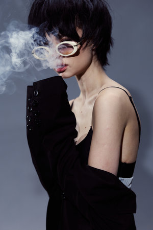 Asian woman smoking with stylish haircut and white sunglasses, wearing black off-shoulder jacket, surrounded by smoke, dark background, moody and mysterious atmosphere.の写真素材