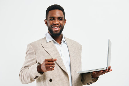 Smiling man with dark skin in business casual outfit holding laptop, gesturing confidently, on white background. Studio portrait of a cheerful professional male demonstrating success and positivity in a modern office setting. Concept: business successの写真素材