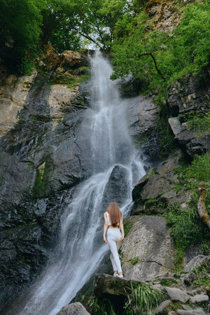 Woman standing near tall waterfall in lush green forest, wearing white outfit and enjoying natures beauty surrounded by rocks and flowing water.の写真素材