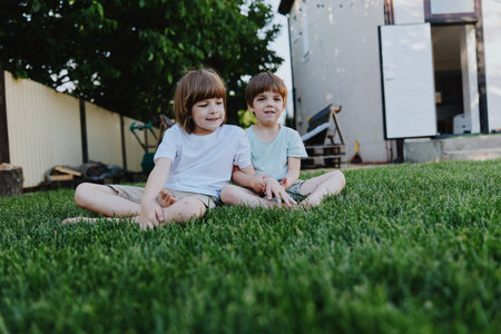 Two cheerful children sitting on green grass, enjoying a sunny day. Their playful expressions capture the essence of childhood joy and innocence.の写真素材