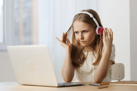 A young girl wearing headphones, engaged in an online lesson, displaying concentration and determination in a bright, modern workspace with a laptopの写真素材