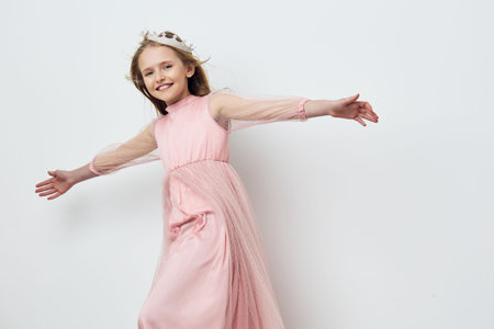 A joyful young girl in a pink dress with flowing sleeves, posing playfully against a minimalist...