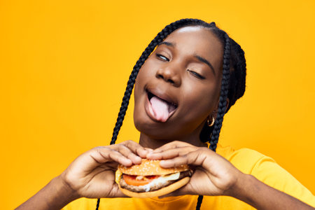 Young woman playfully enjoying a burger against a vibrant yellow background, showcasing fun emotions...