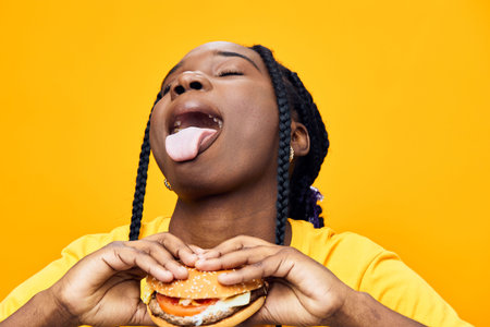 Joyful young woman with braided hair savoring a delicious burger against a vibrant yellow background, expressing excitement and love for fast foodの写真素材