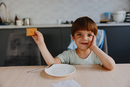 Happy boy holding a cookie, smiling brightly, enjoying a snack at home, showcasing a joyful and playful atmosphere with a cozy kitchen background.の写真素材