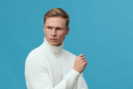 serious young caucasian man wear white knit turtleneck sweater look away thoughtful pose in studio against blue background mental health and gadgets conceptの写真素材