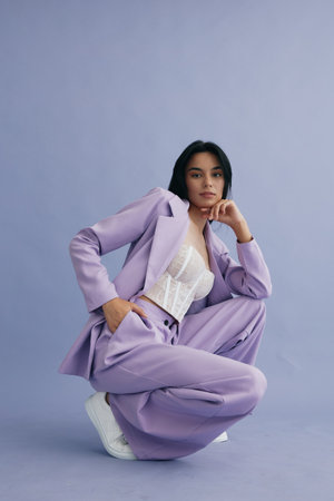 Stylish woman in a lilac pantsuit and white lace top posing confidently against a soft purple background, showcasing modern fashion and casual elegance.の写真素材