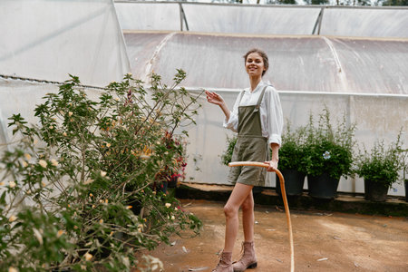 Young woman in casual overalls smiling while watering plants in a bright greenhouse, showcasing...