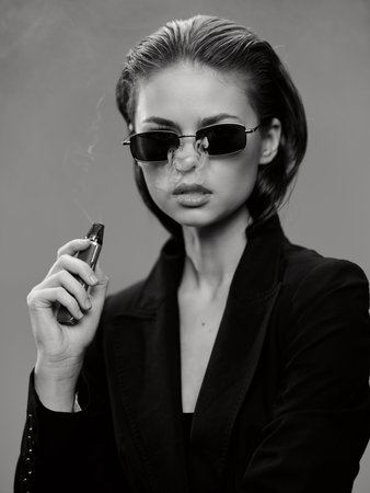 Black and white portrait of woman wearing sunglasses and dark jacket, holding a vape with smoke, showcasing modern style and confident attitude in a minimalistic studio setting.の写真素材