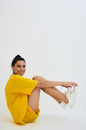 Smiling woman in yellow oversized T-shirt and white sneakers sitting on floor against white background with relaxed pose, casual style, and cheerful expression.の写真素材