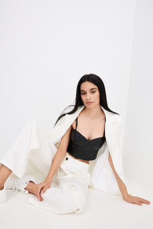 Young woman wearing stylish black bustier and white suit jacket with wide pants sits relaxed on floor against plain white background, showcasing modern fashion.の写真素材