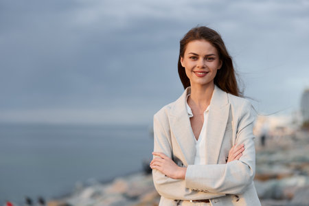 confident woman in light blazer standing outdoors with arms crossed by the sea on cloudy day, natural smile, casual business fashion styleの写真素材