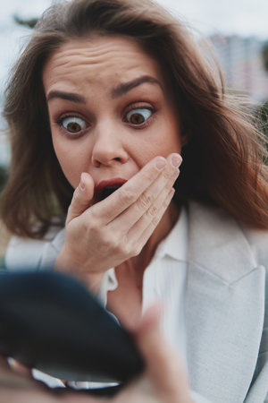shocked woman with wide eyes covering mouth holding smartphone outdoors in business style clothes emotional financial crisis conceptの写真素材