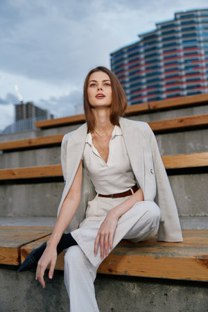Confident woman in stylish white outfit sitting outdoors in urban environment with modern architecture and moody sky, showing elegance and calmの写真素材