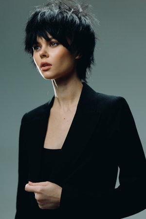 young woman with short black hair wearing a black blazer, looking thoughtfully against a gray studio...