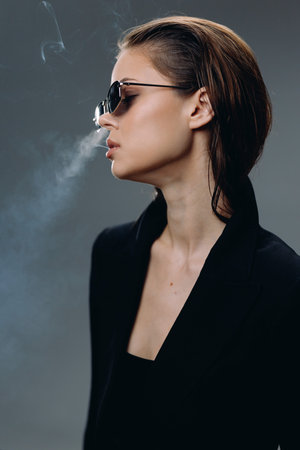 stylish woman in sunglasses smoking with smoke around, wearing black top in moody dark background, artistic portrait of young adultの写真素材