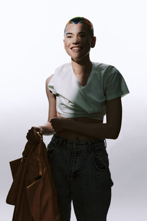 Young woman with short hair smiling, dressed in casual white shirt and jeans, holding a brown jacket, standing against a bright neutral background.の写真素材