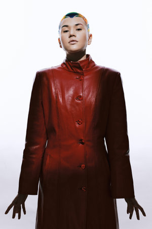 Young woman with short colorful hair wearing a long red leather coat standing confidently against white background with dramatic lighting and modern style.の写真素材