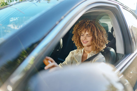 Smiling woman driving car with curly hair in sunny daytime, showing happiness and freedom on a bright urban street.の写真素材