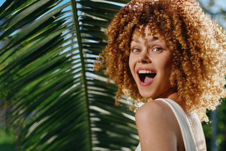 Smiling woman with curly hair enjoys tropical sunlight surrounded by lush green palm leaves, expressing happiness and natural beauty in vibrant outdoor setting.の写真素材
